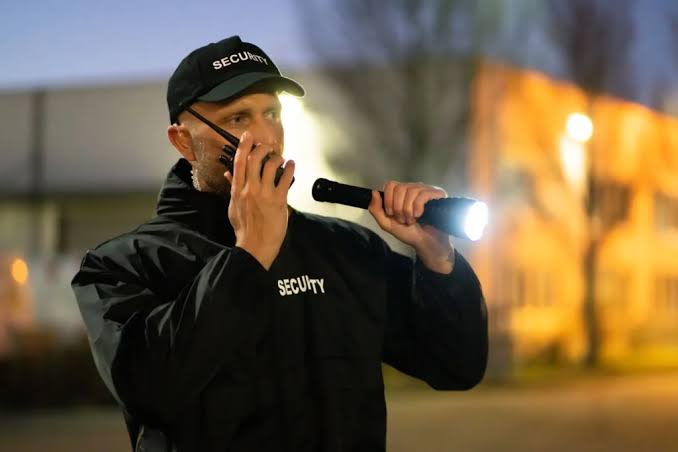 Security officer with a flashlight and radio checking a property perimeter after dark
